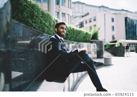 Businessman sitting on bench next to business center and smiling away 65924115