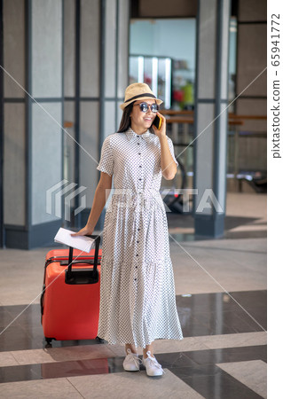 Brunette female in straw hat and sunglasses walking with red suitcase, talking on her smartphone, smiling 65941772