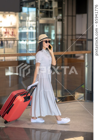 Brunette female in straw hat and sunglasses walking with red suitcase, talking on her smartphone Brunette female in straw hat and sunglasses walking with red suitcase, talking on her smartphone 65941776