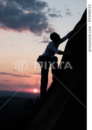 The silhouette of a young strong man climbing a rock wall at sunset. 65946010