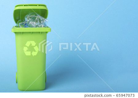 Green trash can with plastic waste and recycling icon on a blue background with place for text 65947073