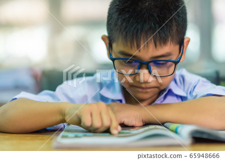 Asian elementary school boy in a white school uniform and wearing glasses, is reading a book in the classroom. 65948666