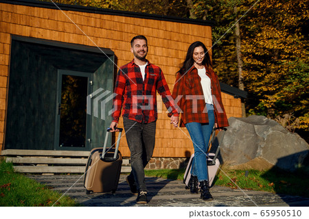 Handsome bearded young man and pretty stylish woman going out from modern house with suitcases while going together to vacation trip. 65950510