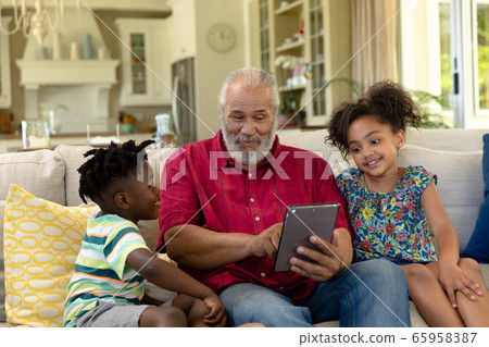 Senior mixed race man sitting on the couch with his young grandson and granddaughter 65958387