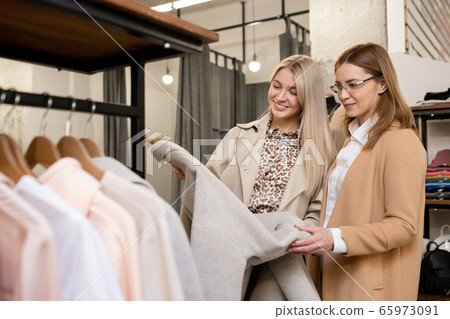 Two pretty females in smart casualwear looking at elegant light grey coat 65973091