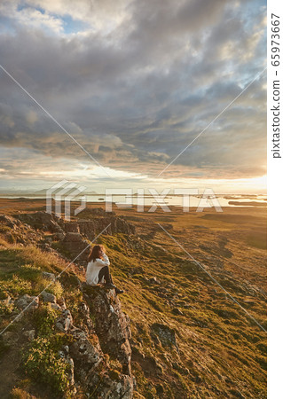 Young woman, female traveler sitting on the top of mountain at sunset. Cloudy dramatic sky. Enjoying wonderful nature in Iceland Young woman, female traveler sitting on the top of mountain at sunset. Cloudy dramatic sky. Enjoying wonderful nature in Iceland 65973667