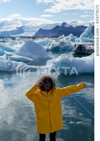 Enjoying Icelandic nature. Excited female tourist in yellow jacket visiting Jokulsarlon glacier lagoon in Iceland. She is looking at camera and smiling Enjoying Icelandic nature. Excited female tourist in yellow jacket visiting Jokulsarlon glacier lagoon in Iceland. She is looking at camera and smiling 65973672