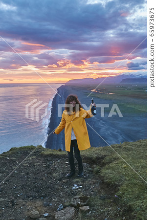 Celebrating Life. Young woman holding a bottle of champagne while admiring the sunset, panoramic view of ocean from the hill in Iceland Celebrating Life. Young woman holding a bottle of champagne while admiring the sunset, panoramic view of ocean from the hill in Iceland 65973675