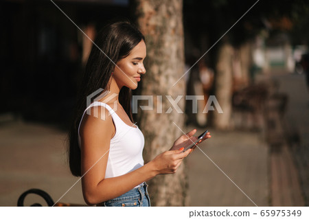 Beautiful girl use tablet outdoors. Female sitting on the bench alone. Woman watch video on tablet and smile. Space for text 65975349