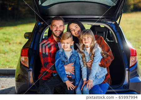 Appealing young modern mother and father and their smiling teen kids which sitting together in the black car's trunk and looking at camera Appealing young modern mother and father and their smiling teen kids which sitting together in the black car's trunk and looking at camera 65997994