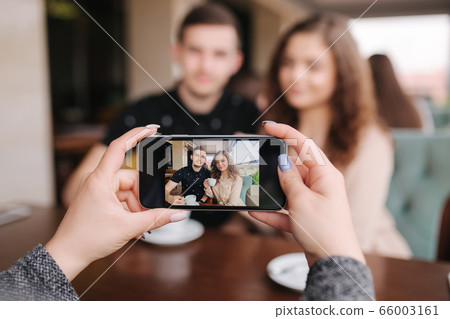 Hand of female making photo of beutiful young couple in cafe. Happy smiled couple posing for photo on smartphone Hand of female making photo of beutiful young couple in cafe. Happy smiled couple posing for photo on smartphone 66003161