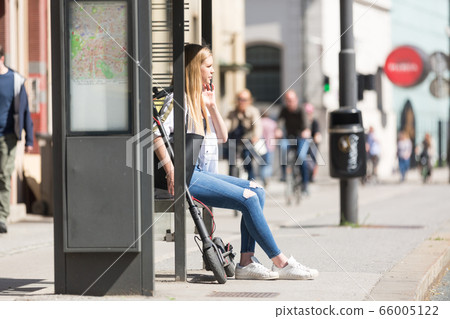 Casual caucasian teenager commuter with modern foldable urban electric scooter sitting on a bus stop bench waiting for metro city bus. Urban mobility concept Casual caucasian teenager commuter with modern foldable urban electric scooter sitting on a bus stop bench waiting for metro city bus. Urban mobility concept 66005122