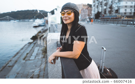 Pretty Asian girl on riverbank. Stylish brunette in a cap with a suitcase resting on river embankment on modern city background. Adventure concept Pretty Asian girl on riverbank. Stylish brunette in a cap with a suitcase resting on river embankment on modern city background. Adventure concept 66007416