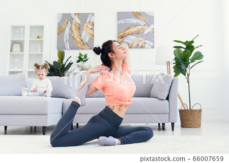 Happy mother doing morning exercises in yoga pose while her little daughter playing at home. Young adorable mom having fun practicing meditation relaxing on stress free weekend with baby girl. 66007659