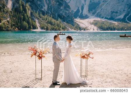 The groom puts on a ring to the bride, at the venue for ceremony, with an arch of autumn flower columns, against the backdrop of Lago di Braies in Italy. Destination wedding in Europe, on Braies lake The groom puts on a ring to the bride, at the venue for ceremony, with an arch of autumn flower columns, against the backdrop of Lago di Braies in Italy. Destination wedding in Europe, on Braies lake 66008043