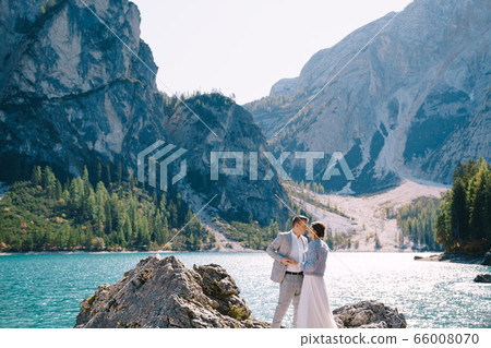 The bride and groom are standing on stones overlooking the Lago di Braies in Italy. Destination wedding in Europe, on Braies lake. Loving newlyweds walk against the backdrop of amazing nature. The bride and groom are standing on stones overlooking the Lago di Braies in Italy. Destination wedding in Europe, on Braies lake. Loving newlyweds walk against the backdrop of amazing nature. 66008070