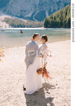 The newlyweds kiss on the spot for the ceremony, with an arch of autumn flower columns, against the backdrop of the Lago di Braies in Italy. Destination wedding in Europe, on Braies lake. The newlyweds kiss on the spot for the ceremony, with an arch of autumn flower columns, against the backdrop of the Lago di Braies in Italy. Destination wedding in Europe, on Braies lake. 66008110