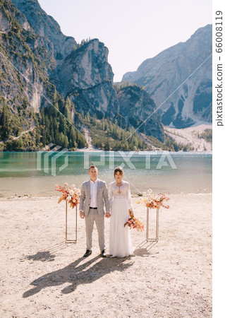 The bride and groom stand in place for the ceremony, with an arch of autumn flower columns, against the backdrop of the Lago di Braies in Italy. Destination wedding in Europe, on Braies lake. The bride and groom stand in place for the ceremony, with an arch of autumn flower columns, against the backdrop of the Lago di Braies in Italy. Destination wedding in Europe, on Braies lake. 66008119