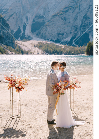 The groom puts on a ring to the bride, at the venue for ceremony, with an arch of autumn flower columns, against the backdrop of Lago di Braies in Italy. Destination wedding in Europe, on Braies lake The groom puts on a ring to the bride, at the venue for ceremony, with an arch of autumn flower columns, against the backdrop of Lago di Braies in Italy. Destination wedding in Europe, on Braies lake 66008123