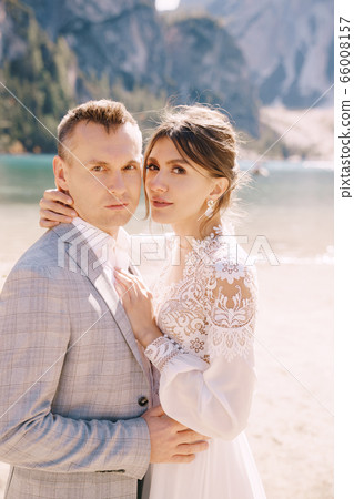 The bride and groom stand in place for the ceremony, with an arch of autumn flower columns, against the backdrop of the Lago di Braies in Italy. Destination wedding in Europe, on Braies lake. The bride and groom stand in place for the ceremony, with an arch of autumn flower columns, against the backdrop of the Lago di Braies in Italy. Destination wedding in Europe, on Braies lake. 66008157