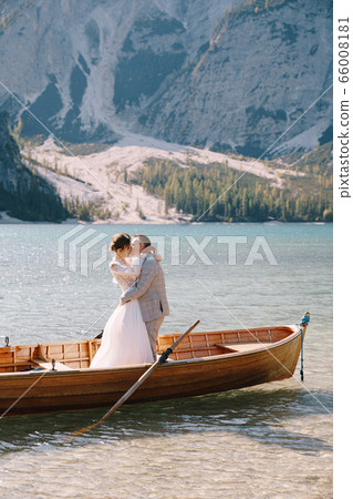 Bride and groom sailing in wooden boat, with oars at Lago di Braies lake in Italy. Wedding in Europe - Newlyweds are standing embracing in a boat. Bride and groom sailing in wooden boat, with oars at Lago di Braies lake in Italy. Wedding in Europe - Newlyweds are standing embracing in a boat. 66008181