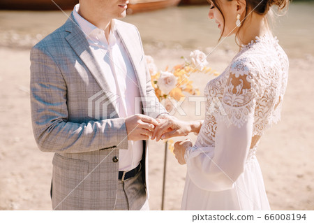 The groom puts on a ring to the bride, at the venue for ceremony, with an arch of autumn flower columns, against the backdrop of Lago di Braies in Italy. Destination wedding in Europe, on Braies lake 66008194