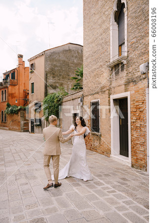 Italy wedding in Venice. The bride and groom walk along the deserted streets of the city. The newlyweds hug, dance, hold hands against the backdrop of picturesque red brick houses. 66008196