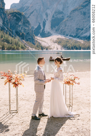 The groom puts on a ring to the bride, at the venue for ceremony, with an arch of autumn flower columns, against the backdrop of Lago di Braies in Italy. Destination wedding in Europe, on Braies lake The groom puts on a ring to the bride, at the venue for ceremony, with an arch of autumn flower columns, against the backdrop of Lago di Braies in Italy. Destination wedding in Europe, on Braies lake 66008212