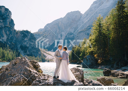 The bride and groom are standing on stones overlooking the Lago di Braies in Italy. Destination wedding in Europe, on Braies lake. Loving newlyweds walk against the backdrop of amazing nature. The bride and groom are standing on stones overlooking the Lago di Braies in Italy. Destination wedding in Europe, on Braies lake. Loving newlyweds walk against the backdrop of amazing nature. 66008214