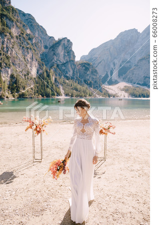 Beautiful bride in a white dress with sleeves and lace, with a yellow autumn bouquet on background of the arch for ceremony, at Lago di Braies in Italy. Destination wedding in Europe, on Braies lake. Beautiful bride in a white dress with sleeves and lace, with a yellow autumn bouquet on background of the arch for ceremony, at Lago di Braies in Italy. Destination wedding in Europe, on Braies lake. 66008273