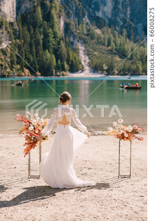 Beautiful bride in a white dress with sleeves and lace, with a yellow autumn bouquet on background of the arch for ceremony, at Lago di Braies in Italy. Destination wedding in Europe, on Braies lake. 66008275