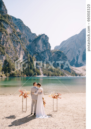 The bride and groom stand in place for the ceremony, with an arch of autumn flower columns, against the backdrop of the Lago di Braies in Italy. Destination wedding in Europe, on Braies lake. The bride and groom stand in place for the ceremony, with an arch of autumn flower columns, against the backdrop of the Lago di Braies in Italy. Destination wedding in Europe, on Braies lake. 66008320