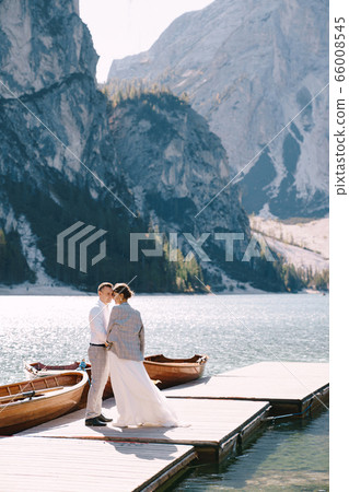 The bride and groom walk along a wooden boat dock at the Lago di Braies in Italy. Wedding in Europe, on Braies lake. Newlyweds walk, kiss, hug on a background of rocky mountains. 66008545