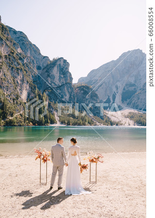 The bride and groom stand in place for the ceremony, with an arch of autumn flower columns, against the backdrop of the Lago di Braies in Italy. Destination wedding in Europe, on Braies lake. The bride and groom stand in place for the ceremony, with an arch of autumn flower columns, against the backdrop of the Lago di Braies in Italy. Destination wedding in Europe, on Braies lake. 66008554