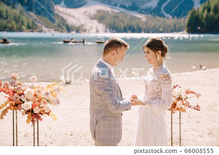 The bride puts a ring on to the groom at venue for the ceremony, with an arch of autumn flower columns, against backdrop of the Lago di Braies in Italy. Destination wedding in Europe, on Braies lake. The bride puts a ring on to the groom at venue for the ceremony, with an arch of autumn flower columns, against backdrop of the Lago di Braies in Italy. Destination wedding in Europe, on Braies lake. 66008555
