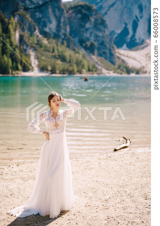Beautiful bride in a white chiffon dress with sleeves and lace on the shore of Lake Lago di Braies in Italy. Destination wedding in Europe, on the popular Braies lake. Beautiful bride in a white chiffon dress with sleeves and lace on the shore of Lake Lago di Braies in Italy. Destination wedding in Europe, on the popular Braies lake. 66008573