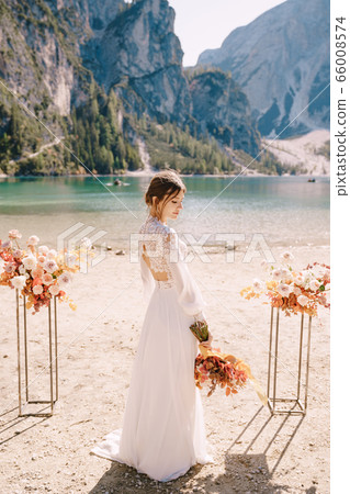 Beautiful bride in a white dress with sleeves and lace, with a yellow autumn bouquet on background of the arch for ceremony, at Lago di Braies in Italy. Destination wedding in Europe, on Braies lake. Beautiful bride in a white dress with sleeves and lace, with a yellow autumn bouquet on background of the arch for ceremony, at Lago di Braies in Italy. Destination wedding in Europe, on Braies lake. 66008574