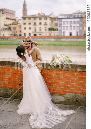 Interracial wedding couple. Wedding in Florence, Italy. African-American bride and Caucasian groom stand embracing on the embankment of the Arno River, overlooking the city and bridges. 66008585