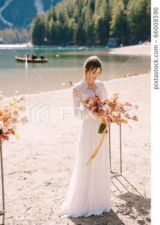 Beautiful bride in a white dress with sleeves and lace, with a yellow autumn bouquet on background of the arch for ceremony, at Lago di Braies in Italy. Destination wedding in Europe, on Braies lake. Beautiful bride in a white dress with sleeves and lace, with a yellow autumn bouquet on background of the arch for ceremony, at Lago di Braies in Italy. Destination wedding in Europe, on Braies lake. 66008590