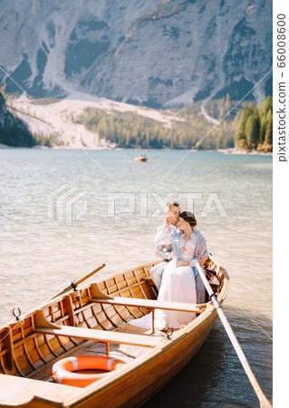 A wedding couple is sitting in a wooden boat on the Lago di Braies in Italy. Newlyweds in Europe, on Braies lake, in the Dolomites. The groom hugs the bride. 66008600