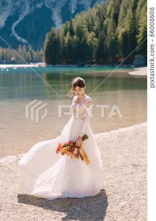 Beautiful bride in a white dress with sleeves and lace, with a yellow autumn bouquet of dried flowers and peony roses, on the Lago di Braies in Italy. Destination wedding in Europe, on Braies lake. Beautiful bride in a white dress with sleeves and lace, with a yellow autumn bouquet of dried flowers and peony roses, on the Lago di Braies in Italy. Destination wedding in Europe, on Braies lake. 66008608