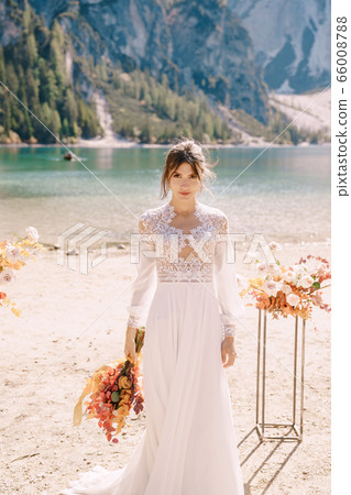 Beautiful bride in a white dress with sleeves and lace, with a yellow autumn bouquet on background of the arch for ceremony, at Lago di Braies in Italy. Destination wedding in Europe, on Braies lake. Beautiful bride in a white dress with sleeves and lace, with a yellow autumn bouquet on background of the arch for ceremony, at Lago di Braies in Italy. Destination wedding in Europe, on Braies lake. 66008788