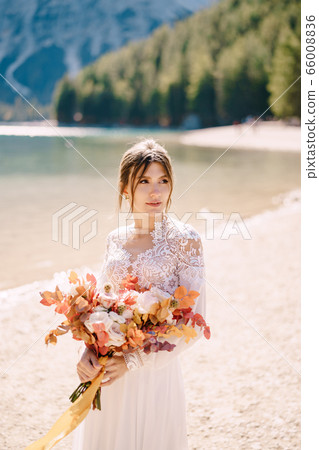 Beautiful bride in a white dress with sleeves and lace, with a yellow autumn bouquet of dried flowers and peony roses, on the Lago di Braies in Italy. Destination wedding in Europe, on Braies lake. Beautiful bride in a white dress with sleeves and lace, with a yellow autumn bouquet of dried flowers and peony roses, on the Lago di Braies in Italy. Destination wedding in Europe, on Braies lake. 66008836