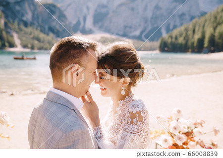 The newlyweds kiss on the spot for the ceremony, with an arch of autumn flower columns, against the backdrop of the Lago di Braies in Italy. Destination wedding in Europe, on Braies lake. The newlyweds kiss on the spot for the ceremony, with an arch of autumn flower columns, against the backdrop of the Lago di Braies in Italy. Destination wedding in Europe, on Braies lake. 66008839
