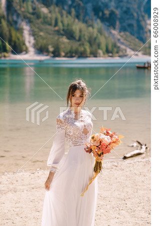 Beautiful bride in a white dress with sleeves and lace, with a yellow autumn bouquet of dried flowers and peony roses, on the Lago di Braies in Italy. Destination wedding in Europe, on Braies lake. 66008929