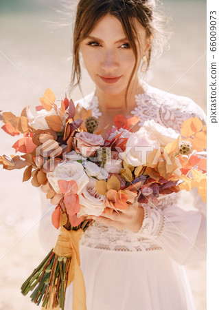 Beautiful bride in a white dress with sleeves and lace, with a yellow autumn bouquet of dried flowers and peony roses, on the Lago di Braies in Italy. Destination wedding in Europe, on Braies lake. Beautiful bride in a white dress with sleeves and lace, with a yellow autumn bouquet of dried flowers and peony roses, on the Lago di Braies in Italy. Destination wedding in Europe, on Braies lake. 66009073
