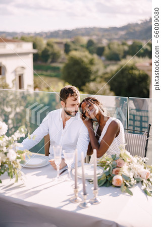 Destination fine-art wedding in Florence, Italy. African-American bride and Caucasian groom are sitting at the rooftop wedding dinner table overlooking the city. Multiracial wedding couple 66009080