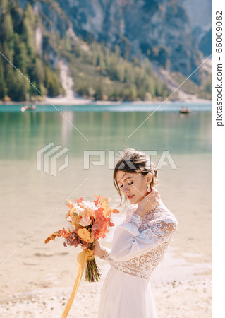 Beautiful bride in a white dress with sleeves and lace, with a yellow autumn bouquet of dried flowers and peony roses, on the Lago di Braies in Italy. Destination wedding in Europe, on Braies lake. 66009082
