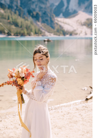 Beautiful bride in a white dress with sleeves and lace, with a yellow autumn bouquet of dried flowers and peony roses, on the Lago di Braies in Italy. Destination wedding in Europe, on Braies lake. Beautiful bride in a white dress with sleeves and lace, with a yellow autumn bouquet of dried flowers and peony roses, on the Lago di Braies in Italy. Destination wedding in Europe, on Braies lake. 66009083