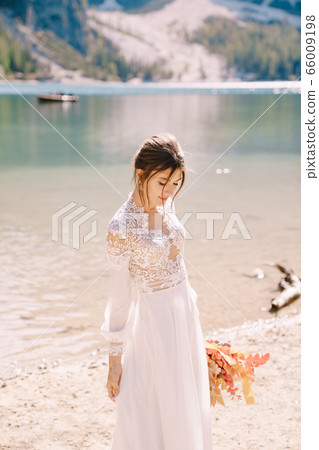 Beautiful bride in a white dress with sleeves and lace, with a yellow autumn bouquet of dried flowers and peony roses, on the Lago di Braies in Italy. Destination wedding in Europe, on Braies lake. 66009198